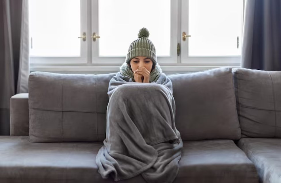 a young woman covered with a blanket wearing a green hat and trying to stay warm while sitting on a grey couch in front of multiple windows