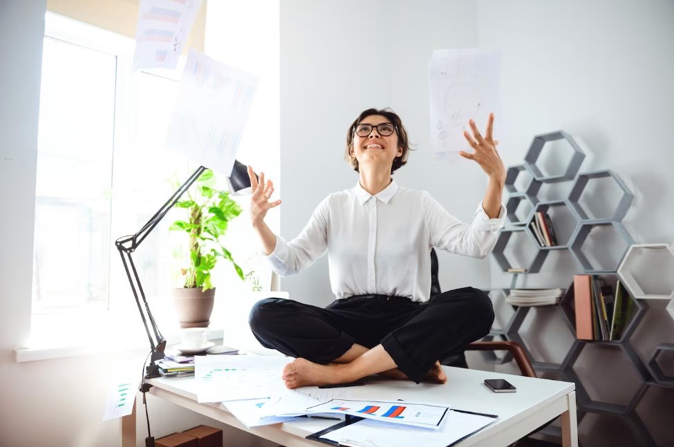 a young businesswoman in glasses and a white shirt smiling and throwing up papers while sitting on a table at the workplace in the office