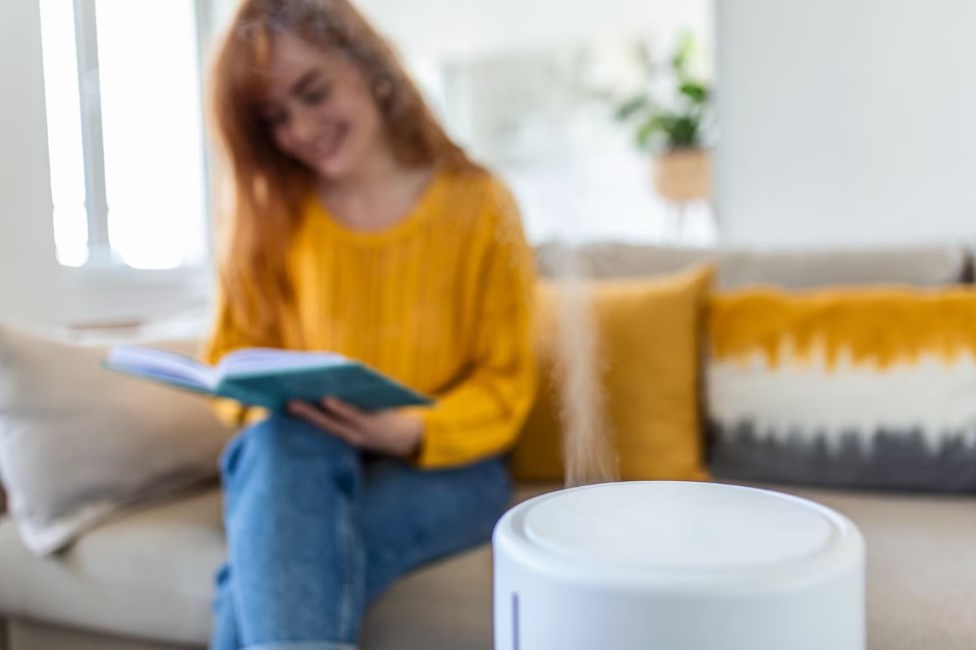 a modern air humidifier and a blurred woman in a yellow sweater resting while reading a book on a sofa in the background