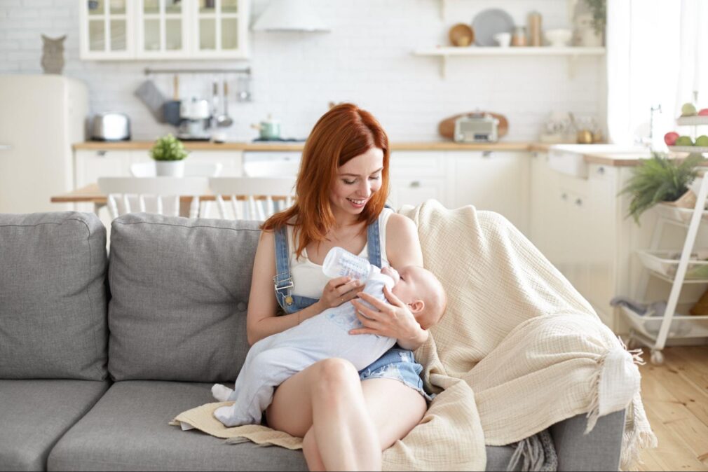 a portrait of a ginger mother bottle-feeding her baby while sitting on a grey couch in the kitchen environment
