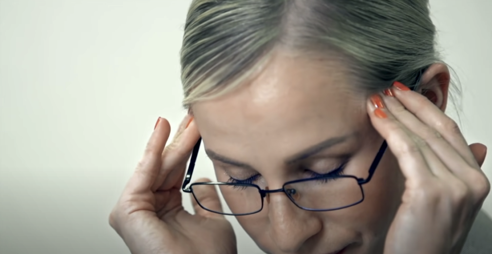 Blonde woman wearing eyeglasses, holding her head on the sides of her forehead with both hands, and closing her eyes.